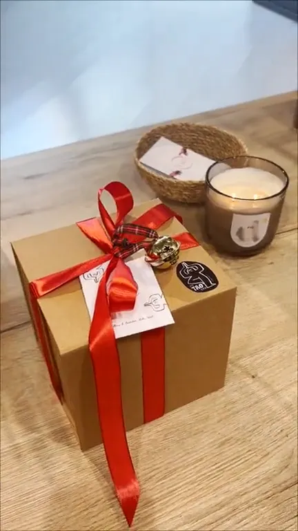 La cajita TAO: caja de cartón kraft con lazo de raso rojo, cascabel, tarjeta y pegatina del logo, junto a una vela